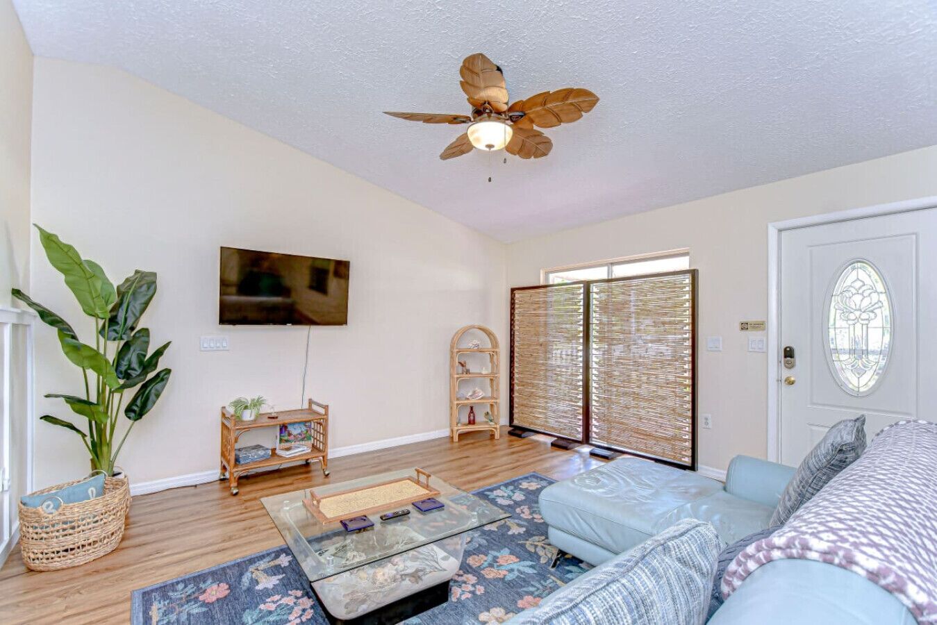 Un salon spacieux doté d’un ventilateur de plafond, d’un téléviseur à écran plat, d’une grande plante, d’une table basse et d’une porte-fenêtre coulissante donnant sur un balcon.