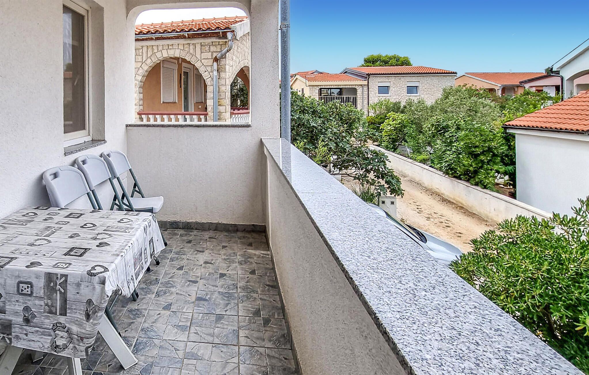 A balcony with a table and chairs, a stone floor, and a view of neighboring houses.