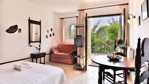 A bedroom with a bed, a sofa, a table with a vase, a TV on a shelf, and a balcony with plants. - 2-Bedroom Holiday Apartment with Beach Access in Cala D'Or (Santanyí)