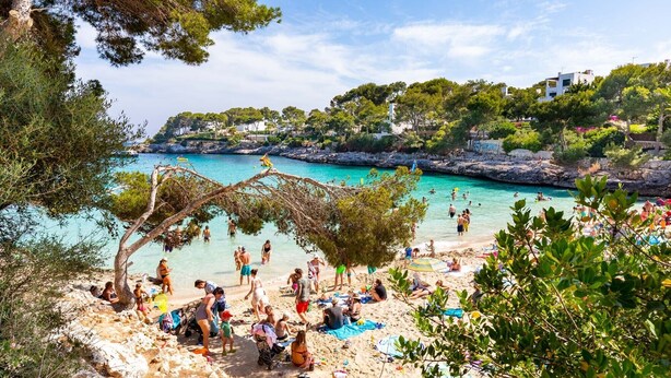 A beach with people sunbathing and swimming, surrounded by greenery and buildings. - 2-Bedroom Holiday Apartment with Beach Access in Cala D'Or (Santanyí)
