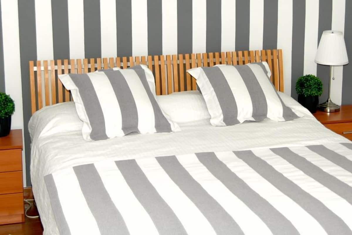 Una cama con colcha a rayas y almohadas, cabecera de madera, mesita de noche con lámpara y una planta pequeña.
