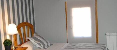 A bedroom with two beds, striped bedding, a wooden headboard, a bedside table with a lamp and a plant, and a window with a view of a white building.