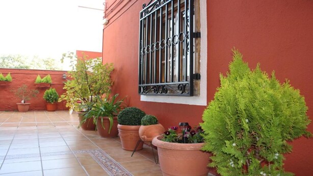 A red building with a window featuring black metal bars, surrounded by potted plants. - Hostal Cervantes (Daganzo de Arriba)