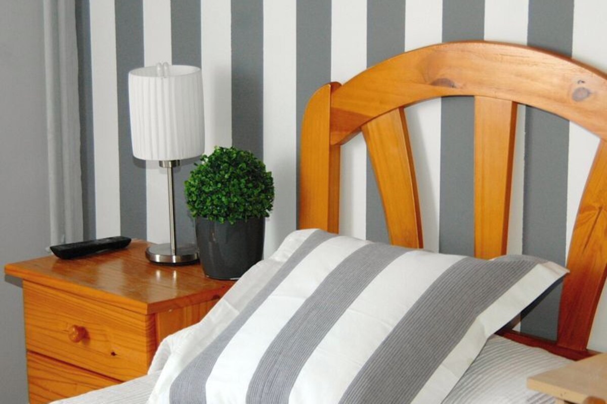 A neatly made bed with a striped pillow, a bedside lamp, a small potted plant, and a wooden nightstand.