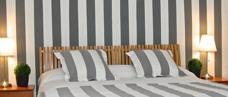 Una cama bien tendida con ropa de cama a rayas, un cabecero de madera y dos mesitas de noche con lámparas y pequeñas macetas.