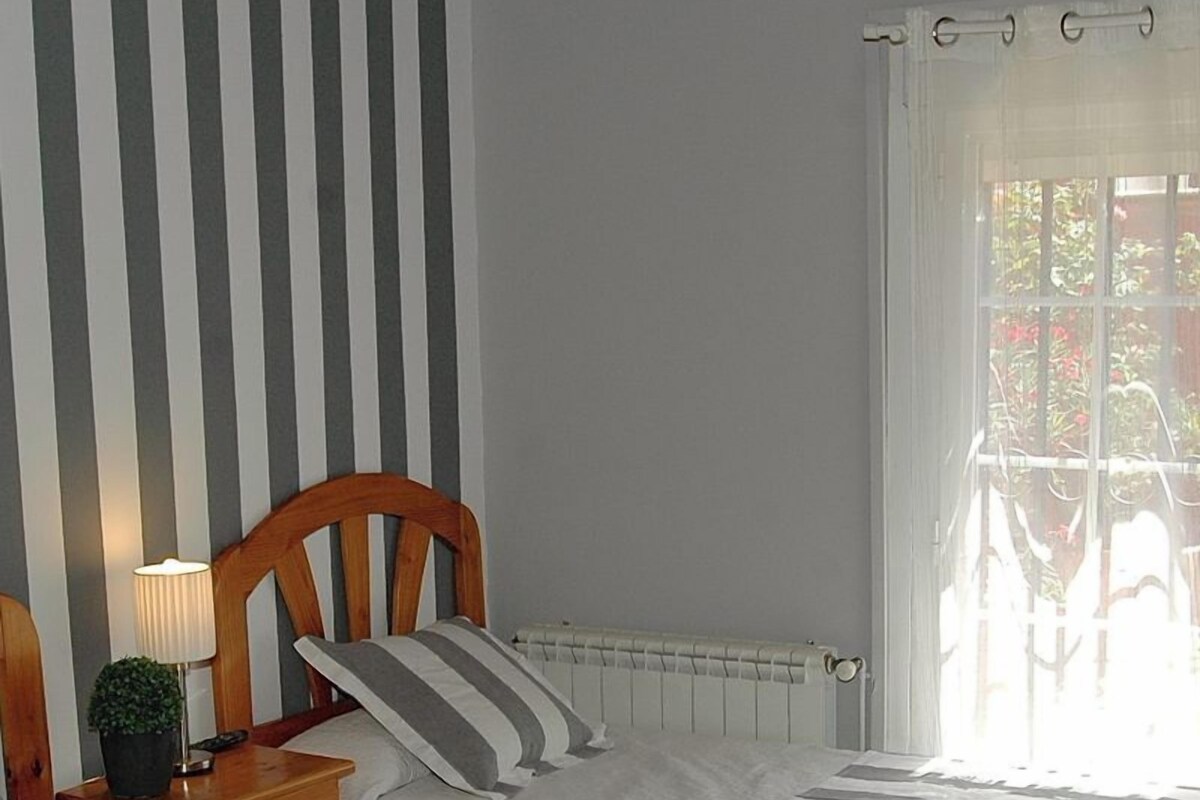 A bedroom with two beds, striped bedding, a nightstand with a lamp, and a window with curtains.