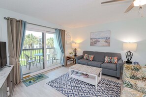 A living room with a sofa, coffee table, and two lamps. There is a sliding glass door leading to an outdoor area with chairs and a table.