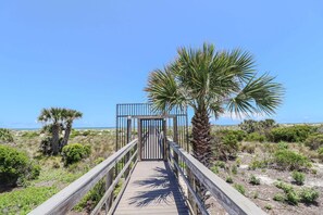 A wooden boardwalk leading to a gated entrance with palm trees and sparse vegetation.