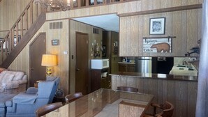 Un vaste espace de vie comprenant une table basse en verre, un canapé et une table à manger. On aperçoit également une cuisine équipée d’un four à micro-ondes et d’un évier. Un escalier mène à l’étage supérieur.