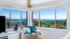 Habitación con un gran ventanal con vistas a un paisaje costero, una zona de estar acogedora y un televisor de pantalla plana sobre un mueble blanco.