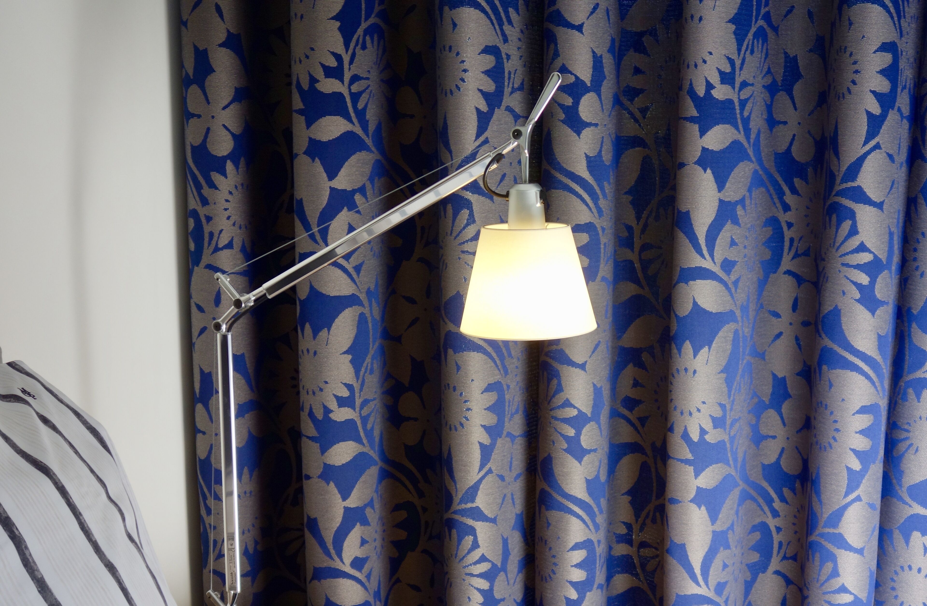 A floor lamp with a white shade and a metal arm, positioned in front of a curtain with a blue floral pattern.