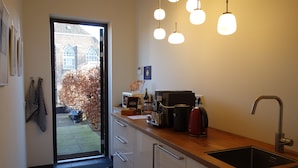 Una cocina moderna con un lavamanos integrado, una cafetera y un hervidor. Hay una ventana con vistas a un edificio de ladrillo y un árbol fuera.