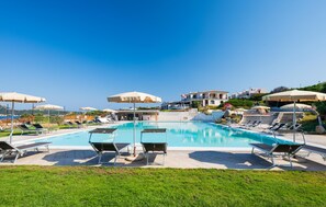 Une piscine entourée de chaises longues et de parasols, avec une pelouse bien entretenue et des bâtiments en arrière-plan.