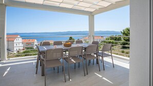 Uma área de refeições coberta ao ar livre, com mesa e cadeiras, com vista para o mar e colinas distantes.