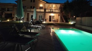 Una piscina con sillas de descanso y sombrillas, un edificio con balcones y un exterior bien iluminado.