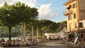 An outdoor café area with tables and chairs, a building with balconies, and a lake in the background. - Casa Bella: Charming Apartment with a Lake View (Bardolino)