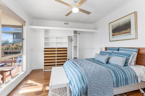 Un dormitorio con cama, ventilador de techo, ventana grande y un cuadro en la pared.
