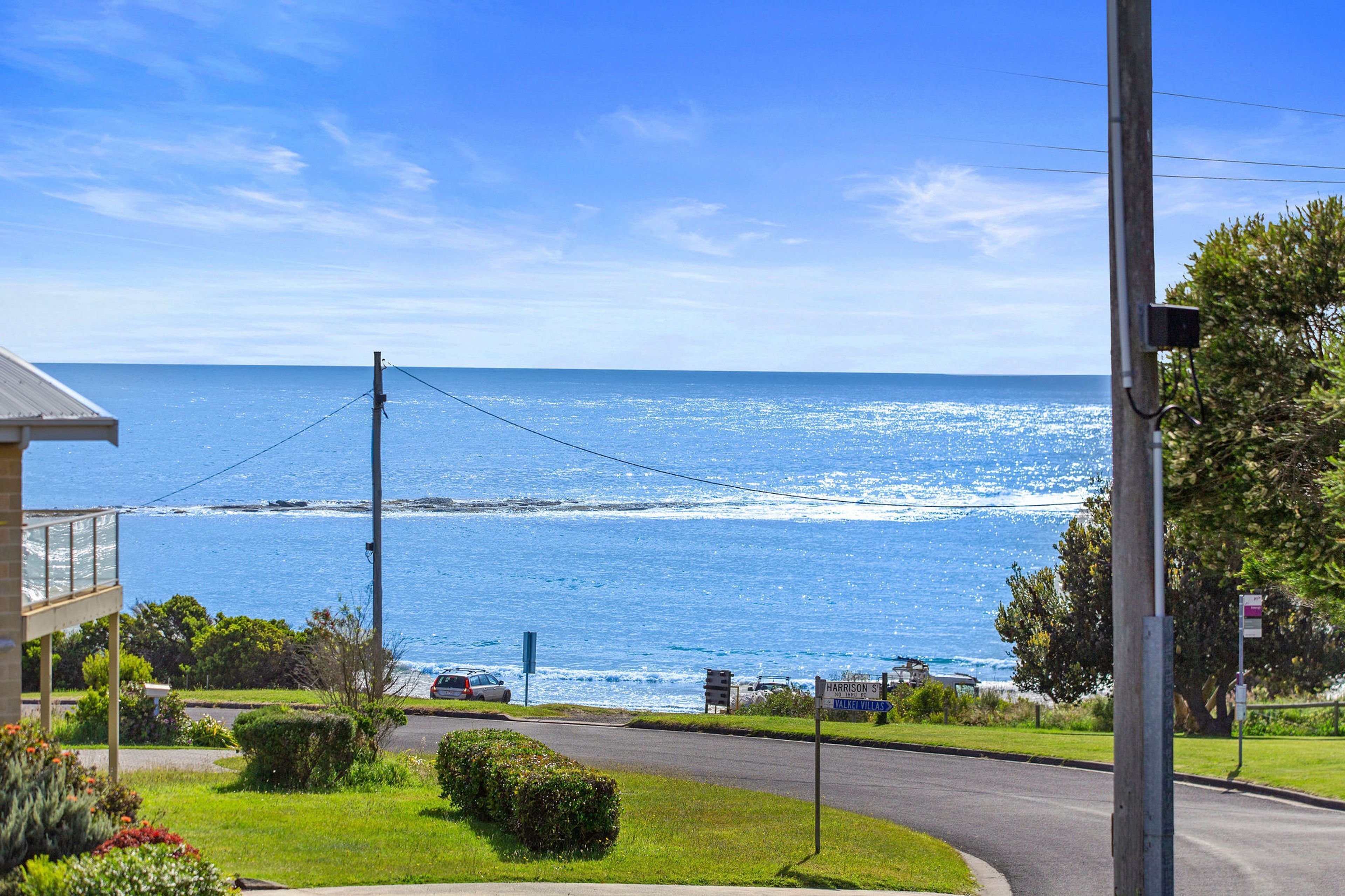 A coastal road with a view of the sea, a clear sky, and a sign indicating 'Harrison St' and 'Vallei Villas'.