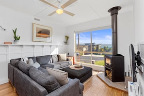 A modern living room with a grey sectional sofa, a fireplace, and a large window offering a view of the outdoors. - Ocean's 8 (Marengo)