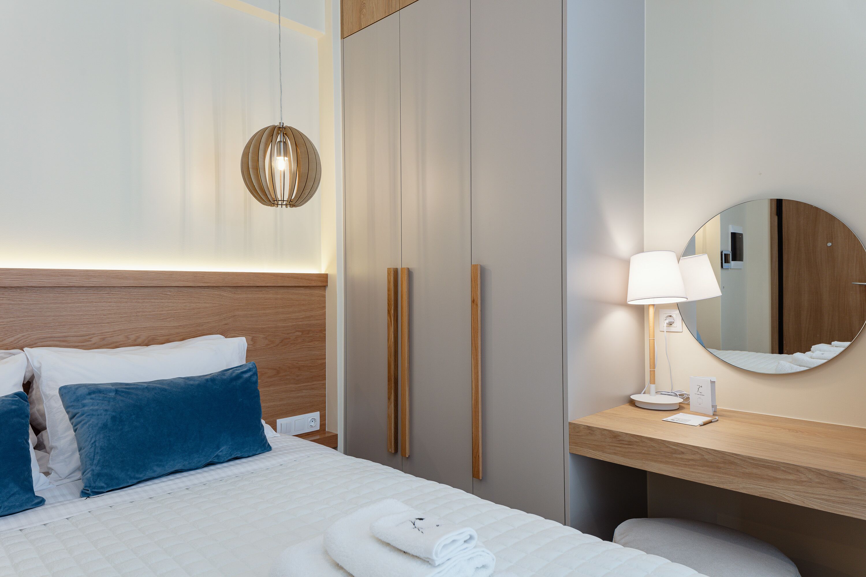 Ein ordentlich bezogenes Bett mit weißen und blauen Kissen, einer Nachttischlampe und einem Spiegel auf einem Holznachttisch.