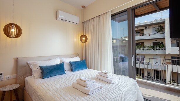 Habitación de hotel con una cama grande, una mesita auxiliar pequeña, un equipo de aire acondicionado y un balcón con vistas a la ciudad.