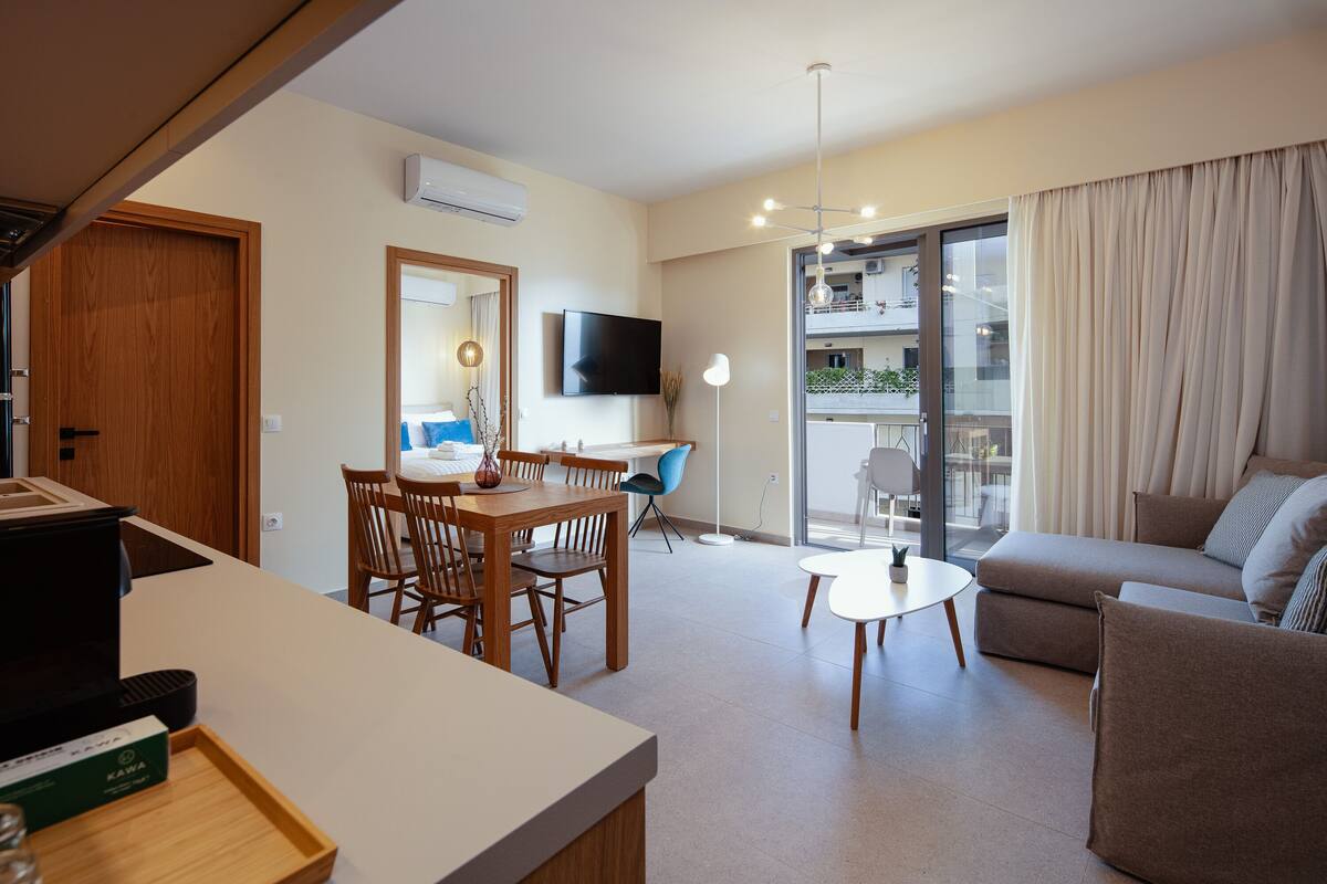 A modern living room with a sofa, a dining table with chairs, a TV mounted on the wall, and a balcony with curtains.