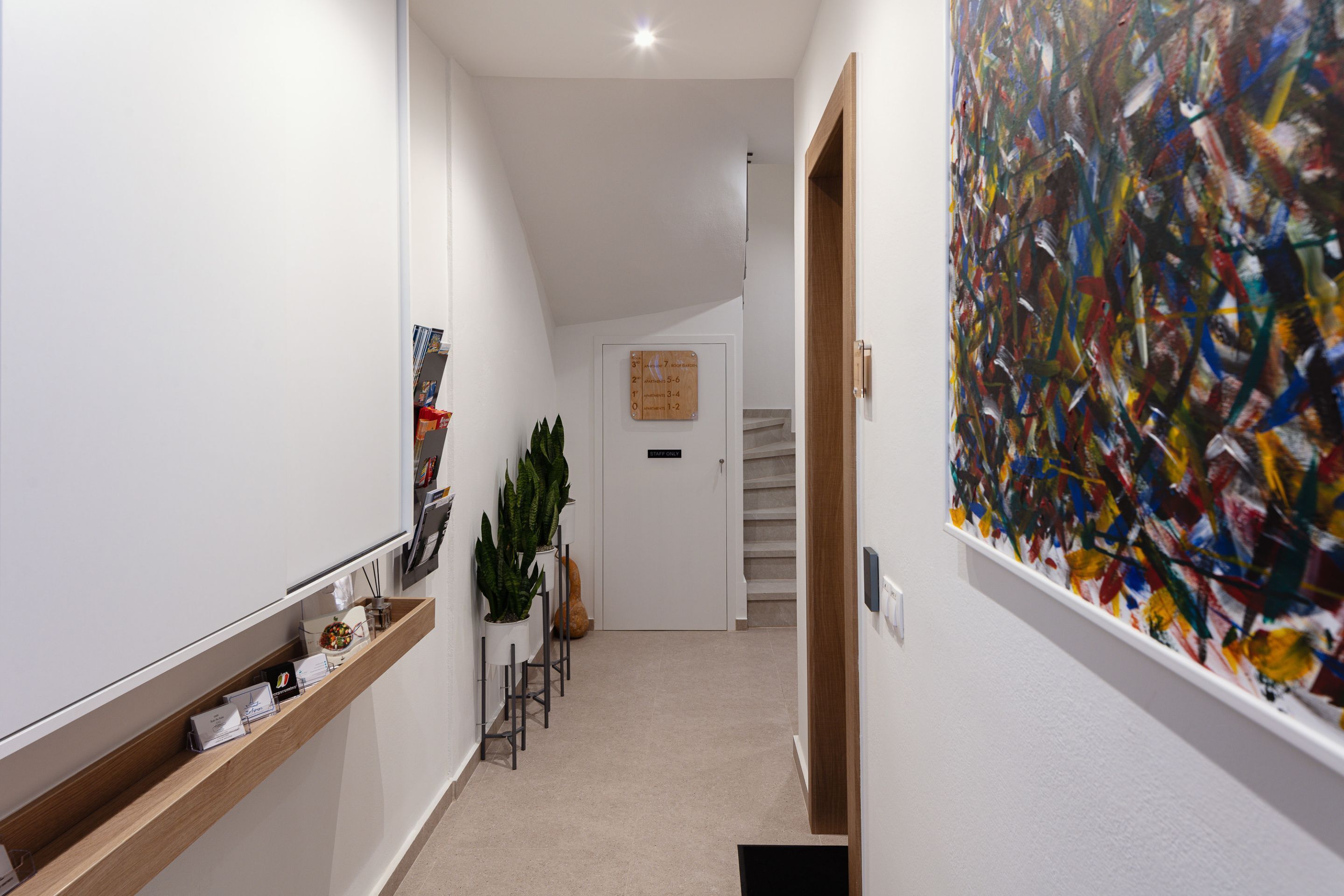 Un couloir avec une étagère en bois, une plante en pot, une œuvre d’art encadrée et un escalier menant à une autre pièce.