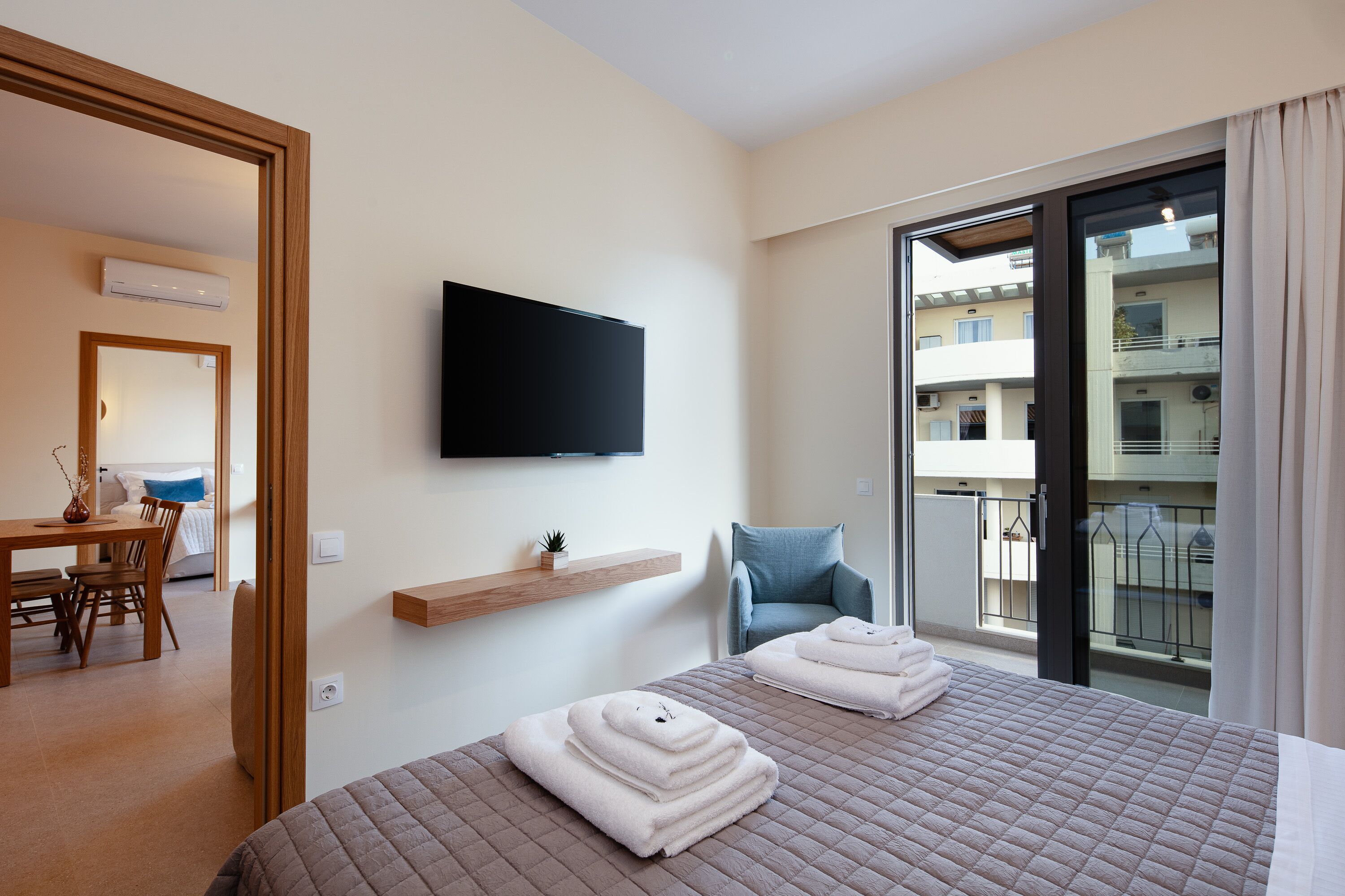 A hotel room with a bed, a TV mounted on the wall, a chair, a small table, and a balcony with a view of the building.