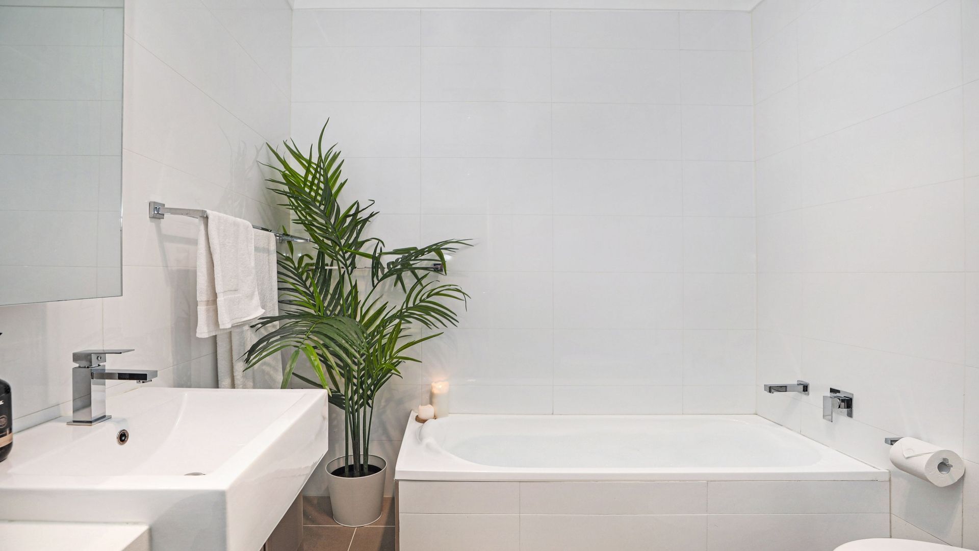 A modern bathroom with a white sink, mirror, and bathtub. There is a potted plant and a candle on the sink.