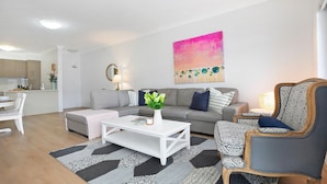 Una sala moderna con un sofá gris, una mesa de centro blanca y un sillón estampado. Se observa una cocina al fondo con encimera blanca y un jarrón con flores.