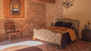 Une chambre avec un mur en pierre, un plafond en bois, un lit avec une literie à motifs, une chaise et une table de chevet avec une lampe.