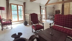 Un salon spacieux avec un canapé, deux fauteuils et une table basse. Une grande fenêtre laisse entrer la lumière naturelle, et on aperçoit un coin cuisine au fond de la pièce.