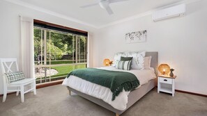 A bedroom with a bed, a chair, a bedside table with a lamp, and a window with a view of greenery.