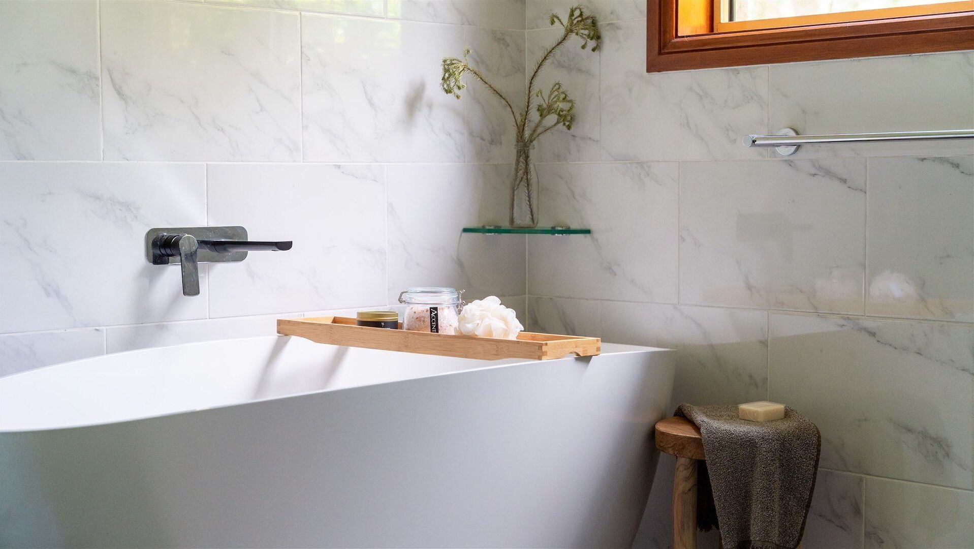 A modern bathroom with a freestanding bathtub, a wooden shelf with toiletries, a glass shelf with decorative plants, and a wooden stool with a towel.
