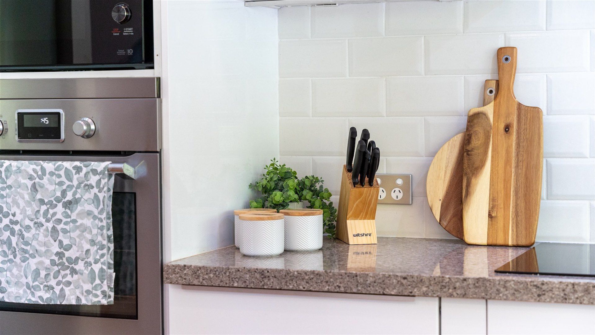 Een keuken met een roestvrijstalen oven, een digitale klok, een houten snijplank, een messenblok en een kleine potplant.