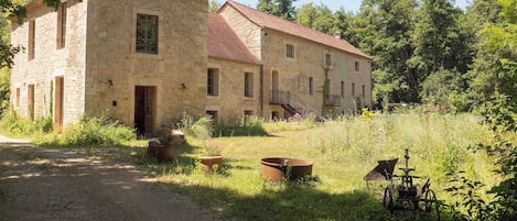 A rustic stone building with a red-tiled roof, surrounded by overgrown grass and trees.