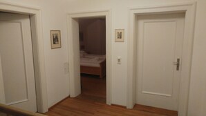 Passadís amb tres portes blanques, una de les quals dona a un dormitori amb un llit i una nitòria. Hi ha quadres amb marc a les parets i un interruptor de llum.
