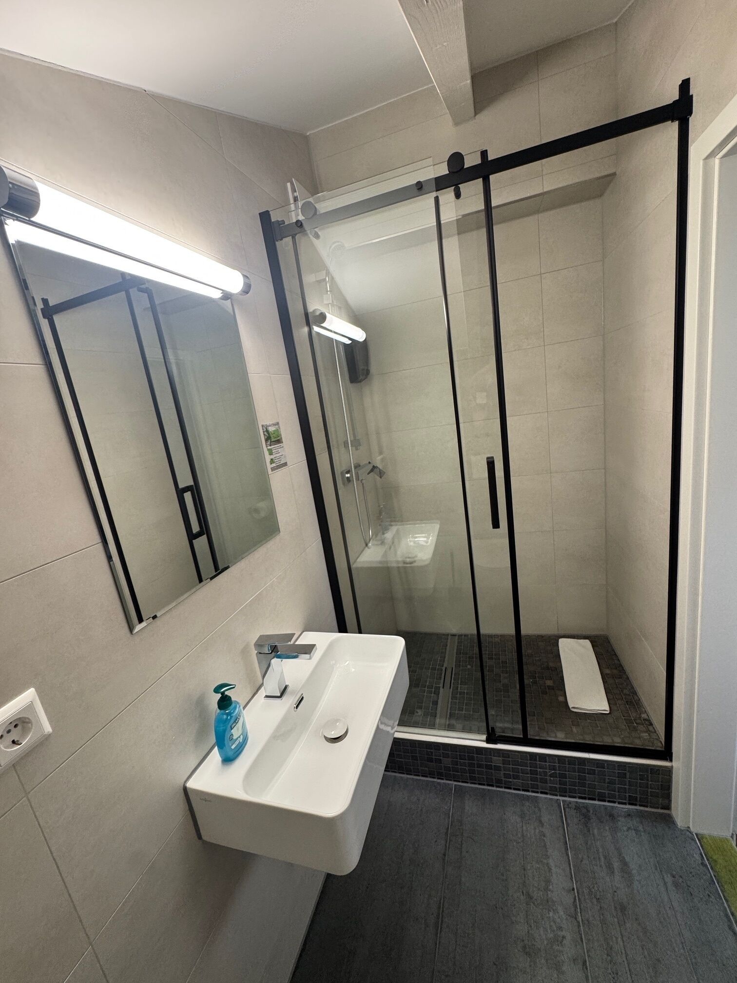 Een moderne badkamer met een douchecabine van glas, een wit wastafelmeubel met zeepdispenser en een spiegel boven de wastafel.