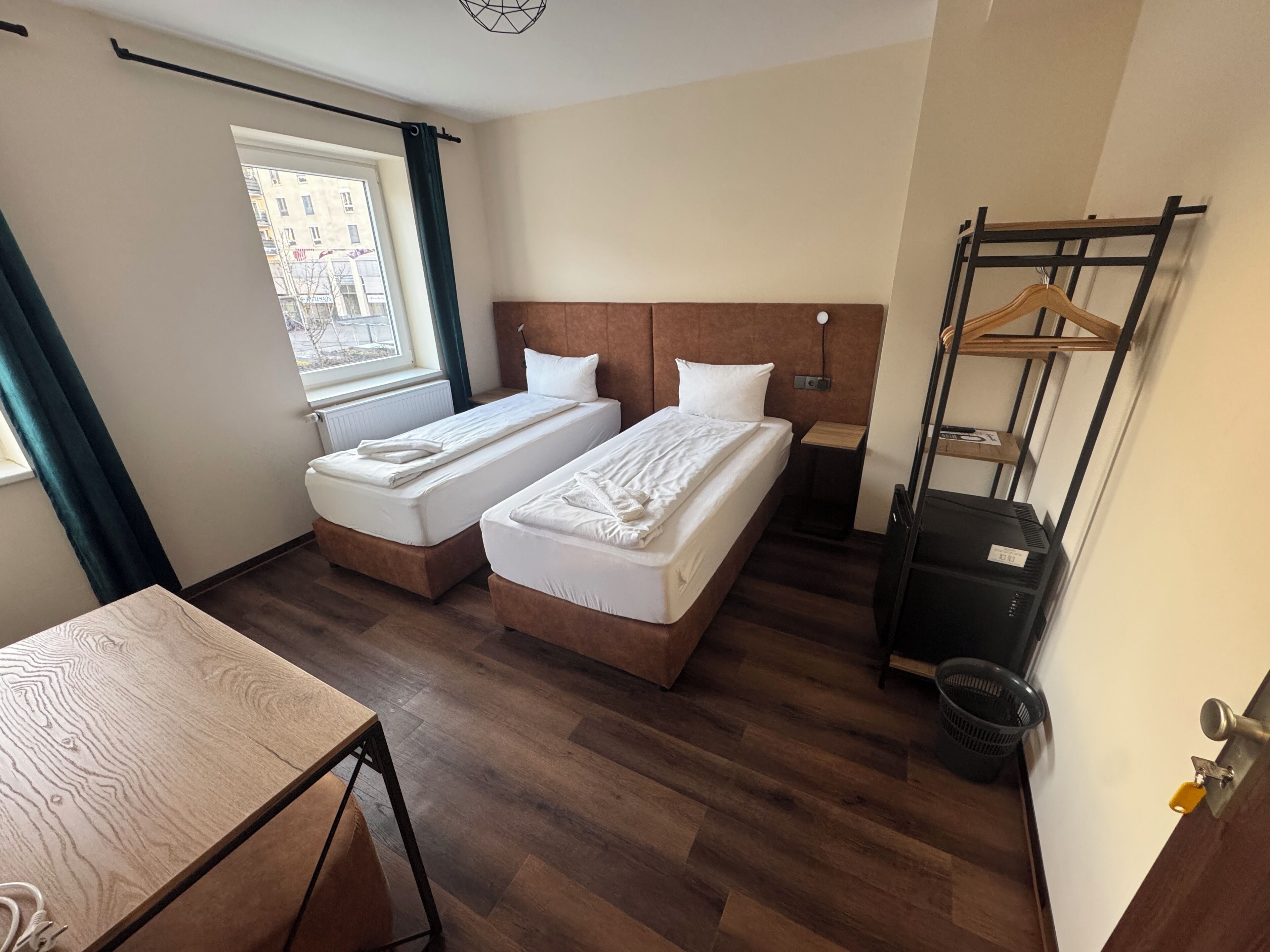 Habitación de hotel con dos camas individuales, una mesa de madera, una ventana con cortinas, un perchero y una maleta negra.