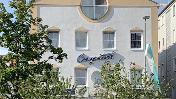 A building with a sign reading "Capitol" and a flag. There are trees and planters in front. - Capitol Zentrum - 24h - (Friedrichshafen)