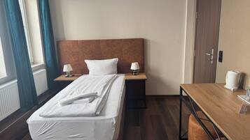 A hotel room with a bed, two bedside tables with lamps, a wooden desk, a chair, a radiator, and a door.