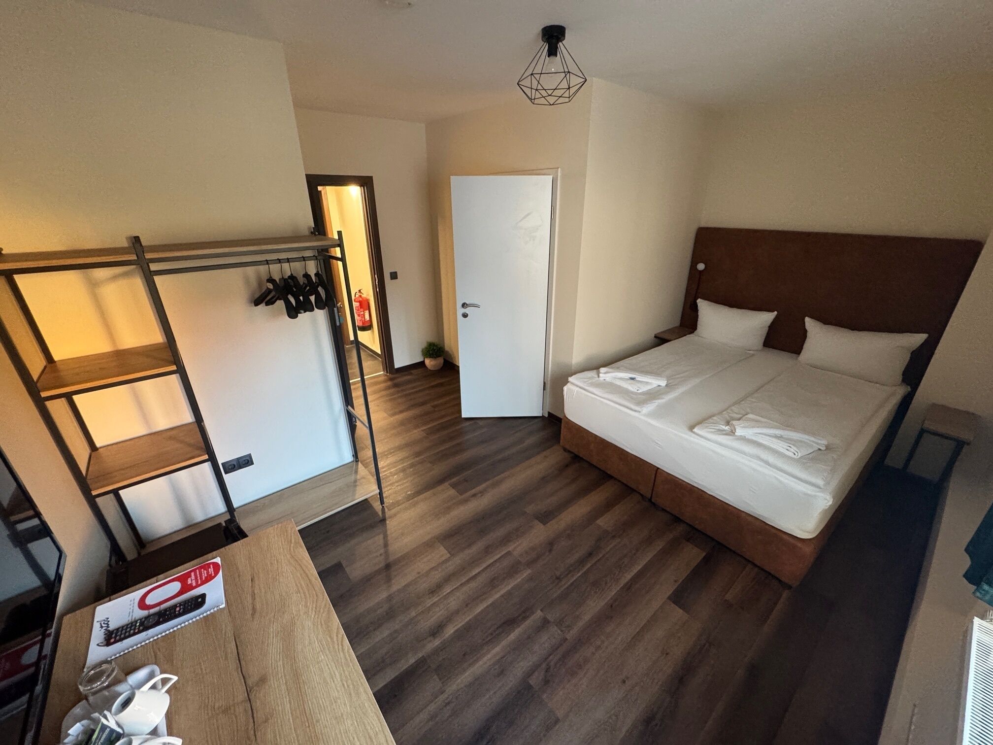 Een hotelkamer met houten vloer, een bed met witte lakens, een houten hoofdbord, een plank met schoenen, een deur en een kleine tafel met een afstandsbediening en brillen.