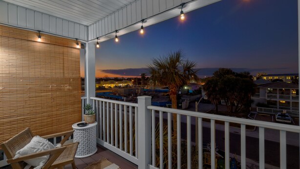 Un balcón con vista a un atardecer, una silla de madera, una mesita con una planta y luces colgantes.