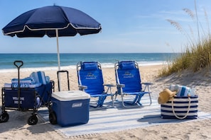 A beach setup with two blue lounge chairs, a Coleman cooler, a beach cart with a 'Welcome Home' sign, and a striped beach bag.
