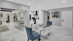 A modern kitchen with white cabinets, a black range hood, and a dining area with a glass table and blue chairs. - LUX 6BR 4Bath Htd Pool & SPA Mini Golf G-Room (Deerfield Beach)