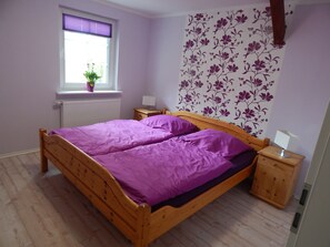 A bedroom with a wooden bed, purple bedding, a window with blinds, a radiator, a nightstand with a lamp, and a floral wallpaper. - Ferienwohnung in zentraler, ländlicher Lage (Groven)
