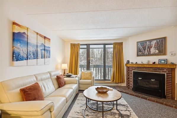 A living room with a sofa, armchair, coffee table, fireplace, and large windows with curtains.