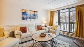 Ein gemütliches Wohnzimmer mit einer Sofagarnitur, einem Couchtisch und einem großen Fenster mit Vorhängen.