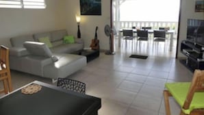 A living room with a sofa, a dining table, and a TV. There is a balcony with chairs and a table. - Villa Alizés 8 pers. 4 air-conditioned bedrooms (Le Gosier)