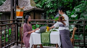 A person in traditional attire serving food to another person at a table with a laptop. - The Sebali Resort (Ubud)